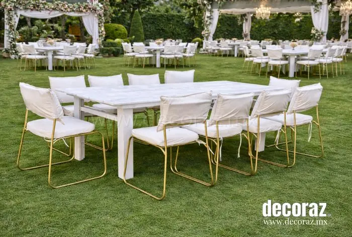 Mesa blanca vintage con sillas metálicas doradas y cojines color marfil para evento en jardín